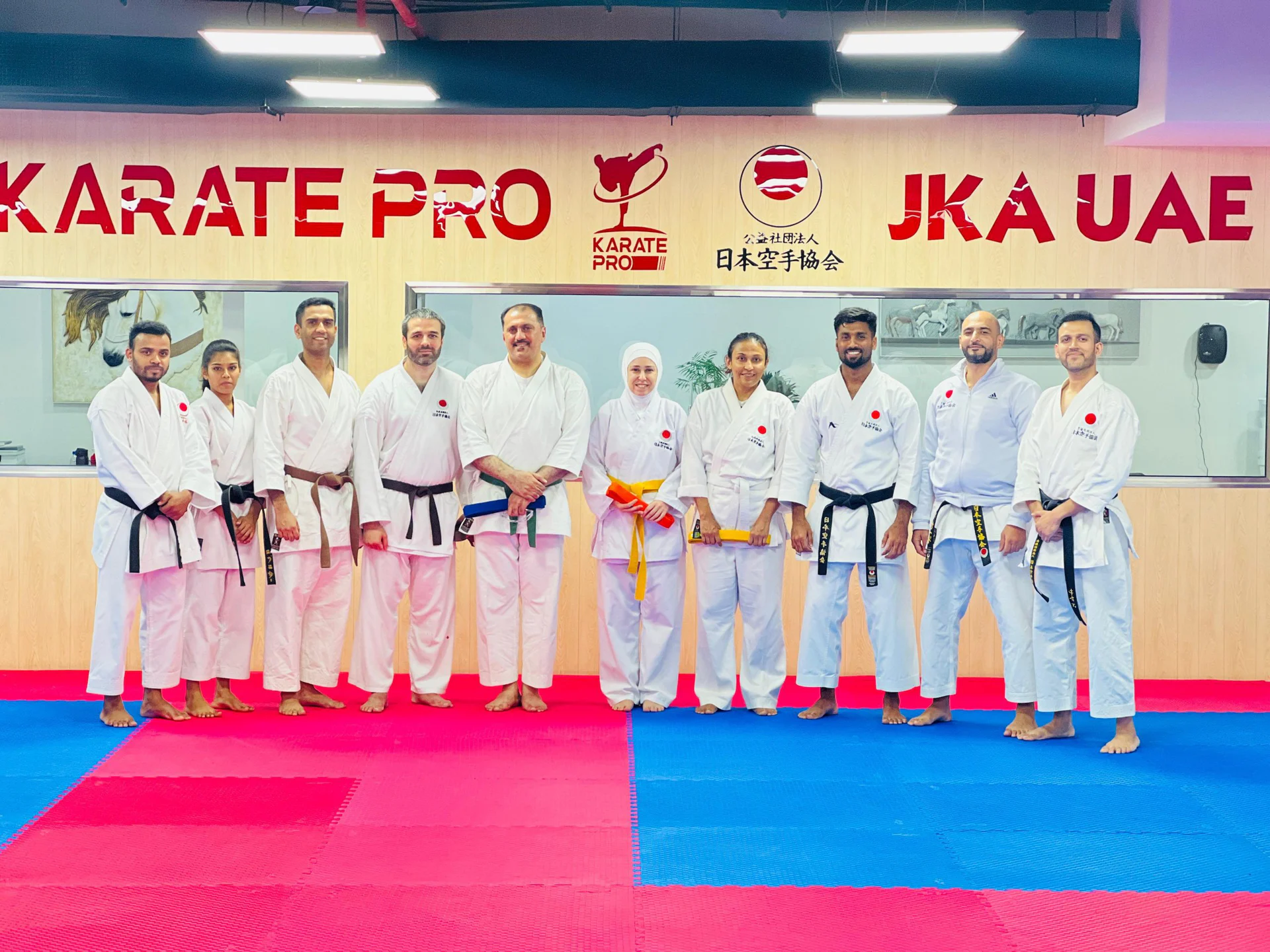 Adult karate class of mixed belt grades posing in a line beneath the Karate Pro and JKA UAE signage