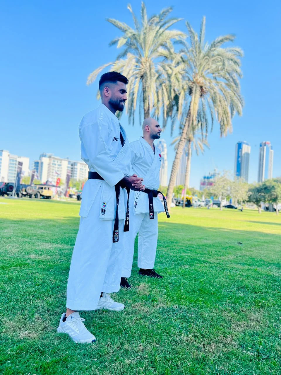Two black belt instructors standing on the grass among palm trees with the Dubai skyline behind them