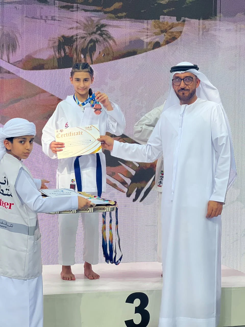 Girl in white gi on the third-place podium receiving her certificate from an official at the 1st International Karate Open Fujairah 2024