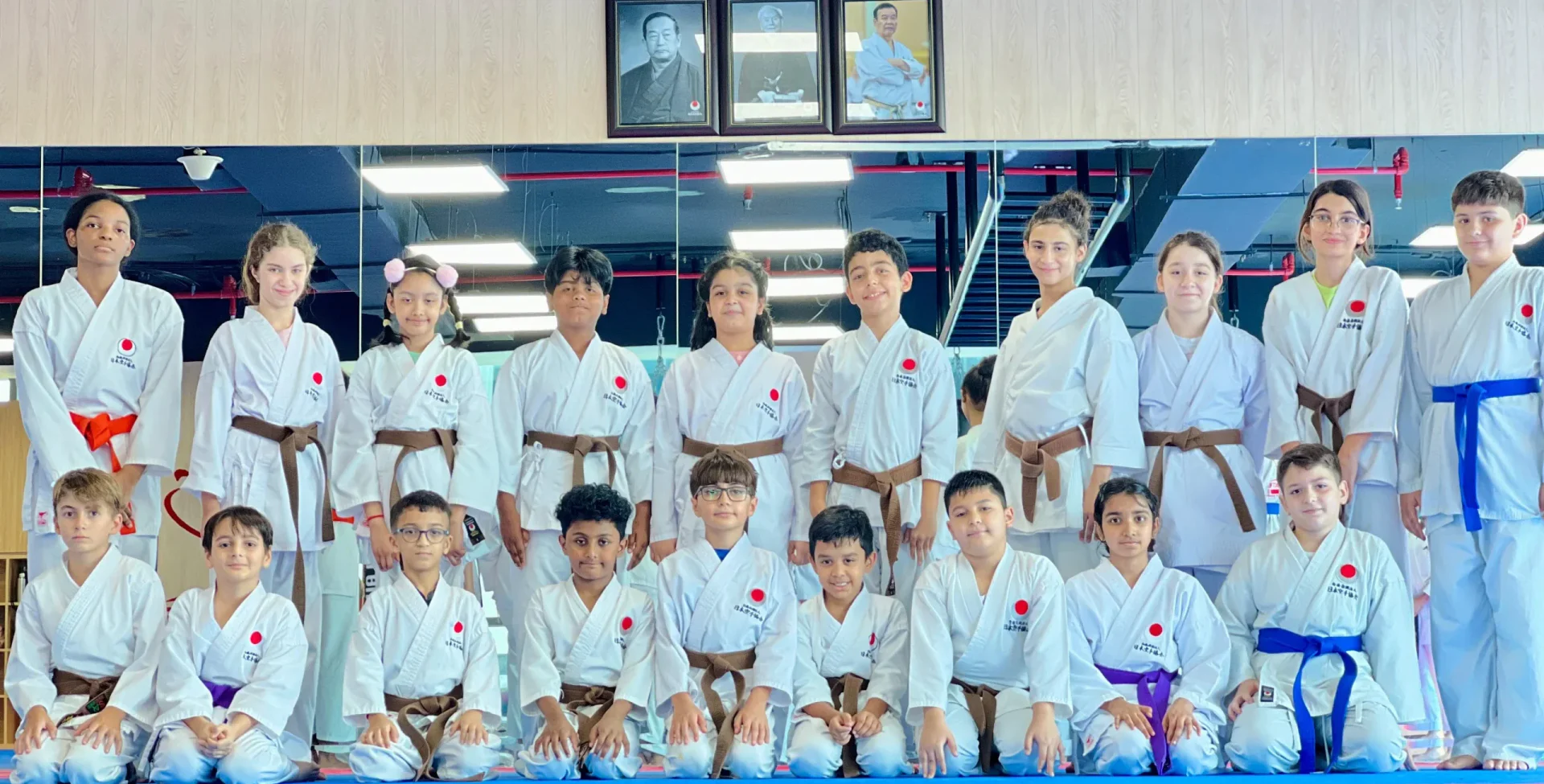 Kids karate class of mixed belt grades posing in rows beneath the dojo's framed portraits