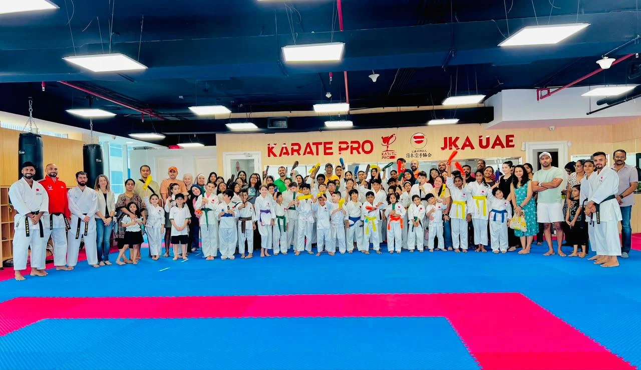 Families and children of mixed belt grades posing together at a belt grading day beneath the Karate Pro and JKA UAE wall signage