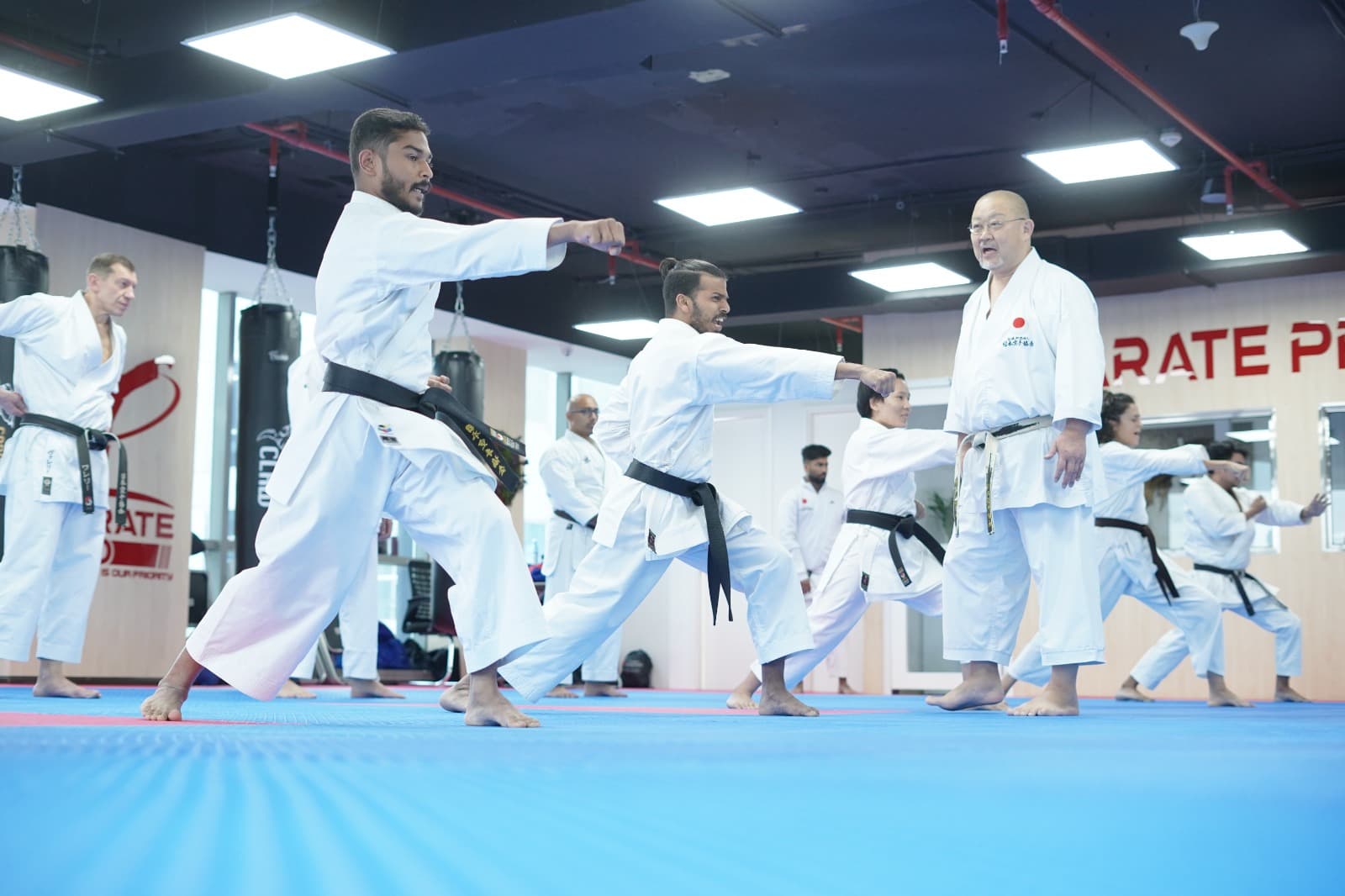 Adult karatekas in white gi practising kata in unison on blue mats, an instructor leading the group in the Karate Pro dojo