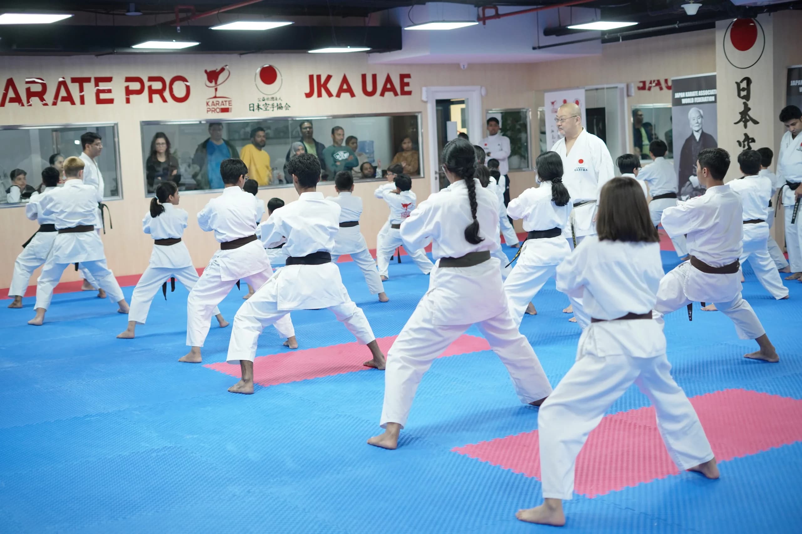 Mixed adult class in white gi holding a deep front stance in rows under the Karate Pro and JKA UAE signs
