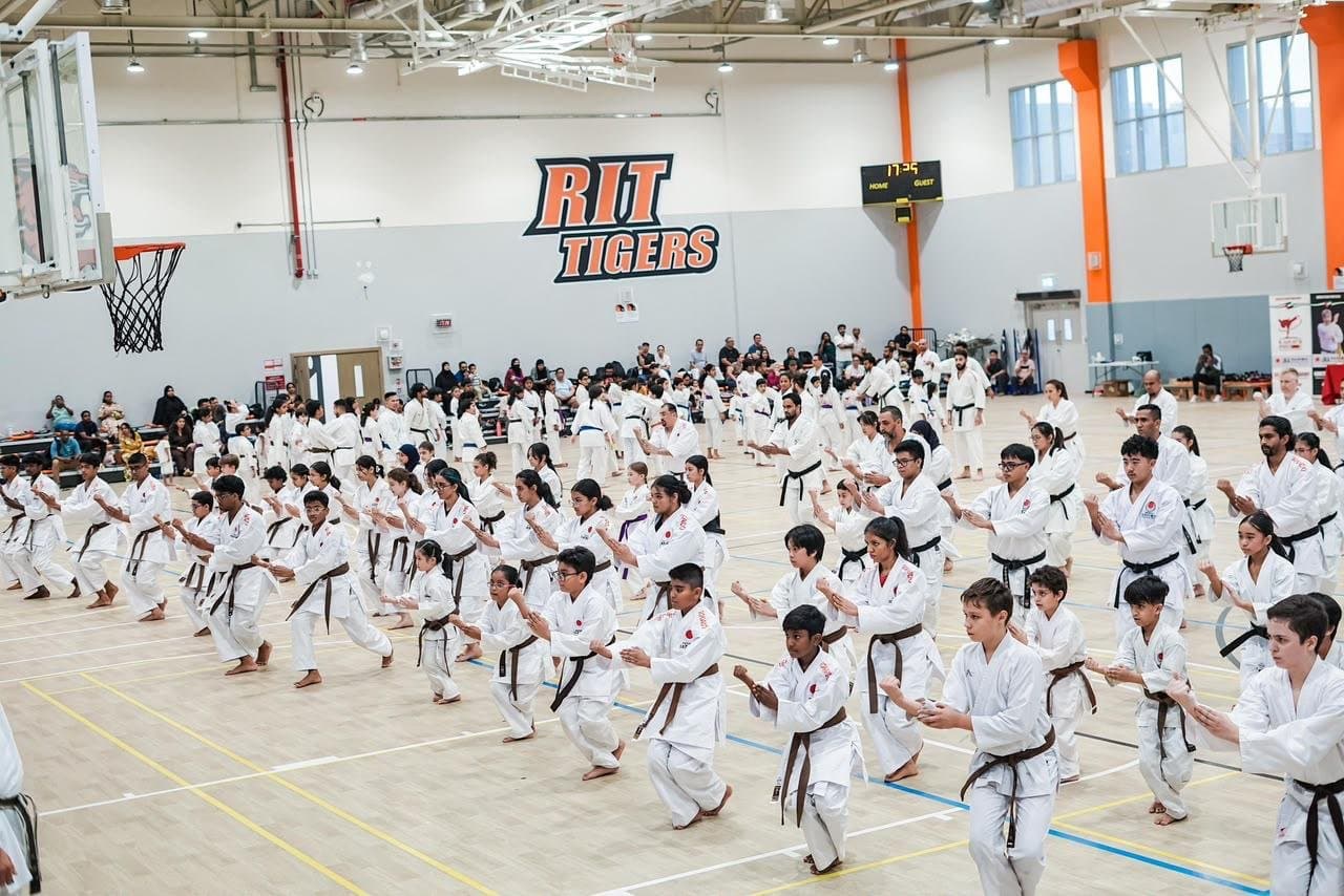 Wide view of a large mixed class practising kata in rows at a JKA Gasshuku seminar beneath the RIT Tigers wall branding