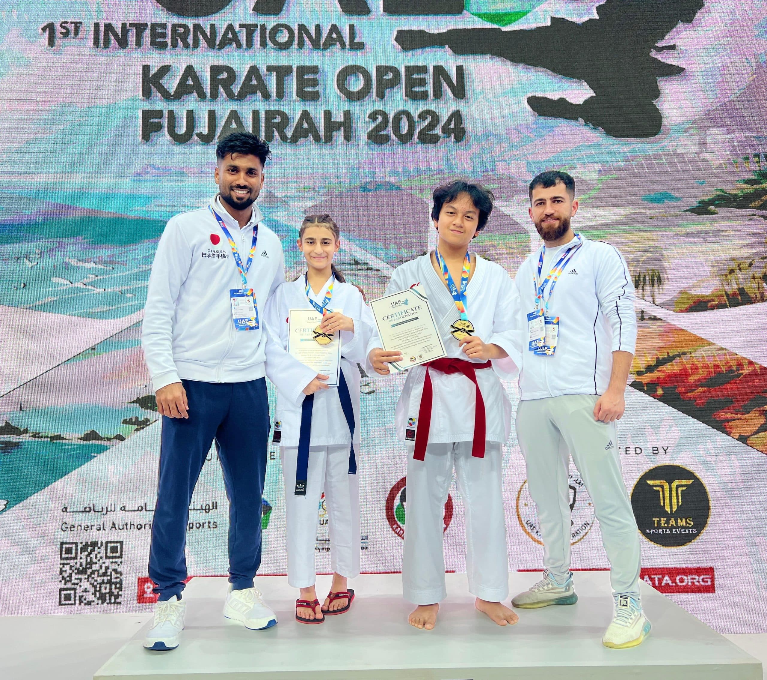 Two young competitors with medals and certificates standing between their coaches at the 1st International Karate Open Fujairah 2024