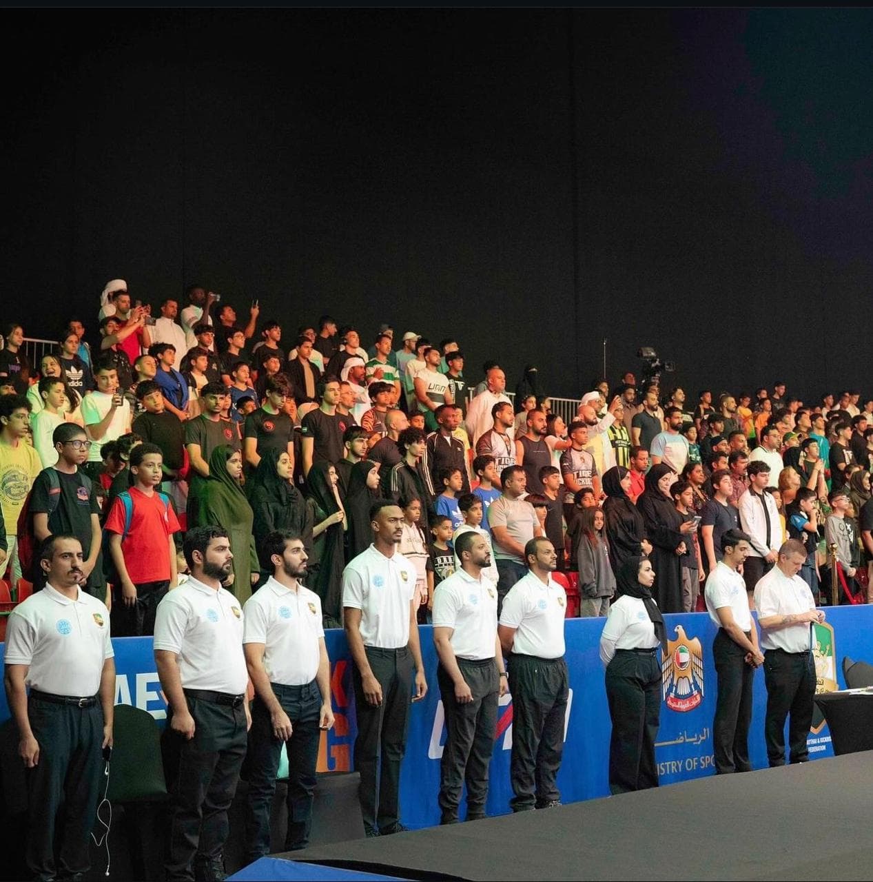 Officials in white shirts standing before a packed crowd of spectators in an indoor arena