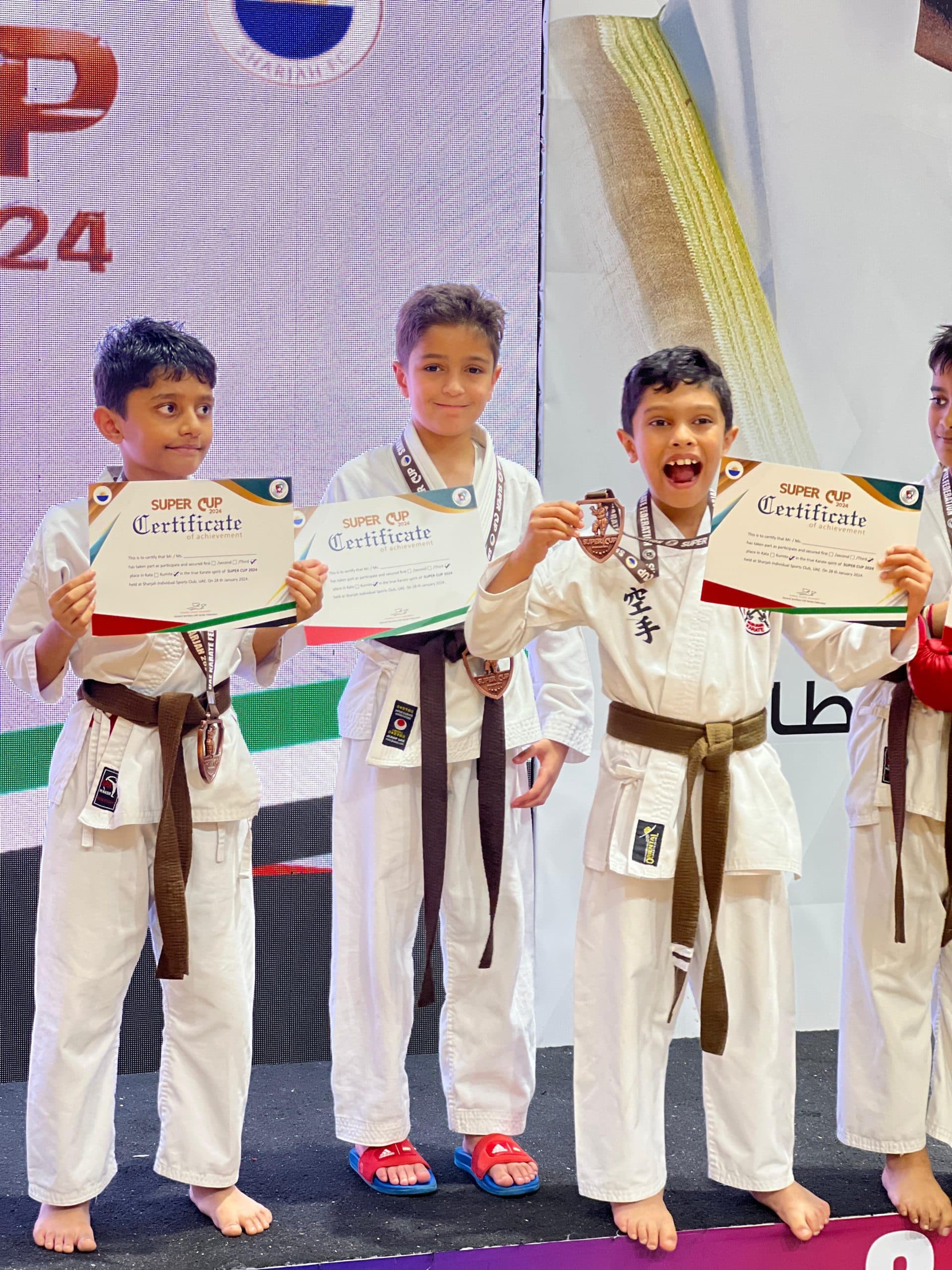 Three boys in white gi proudly holding Super Cup certificates and medals on stage