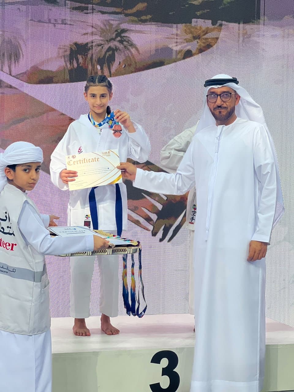 Girl in white gi on the third-place podium receiving her certificate from an official at the 1st International Karate Open Fujairah 2024