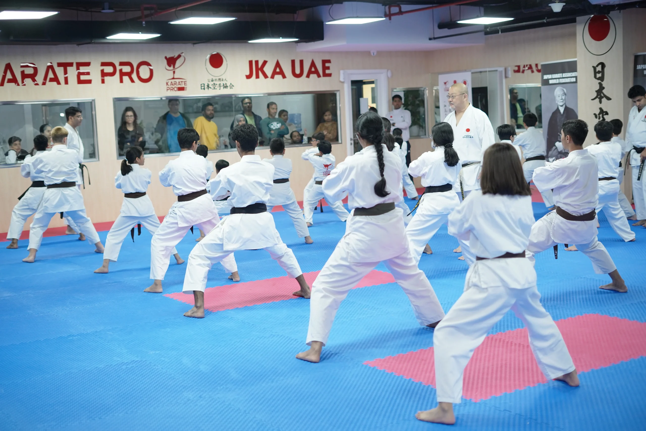 Mixed adult class in white gi holding a deep front stance in rows under the Karate Pro and JKA UAE signs