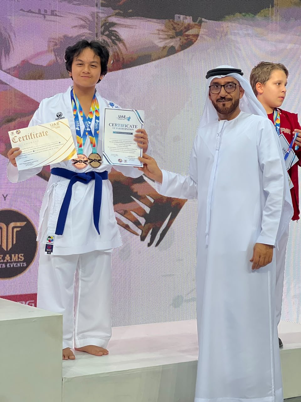 Boy in white gi with a blue belt holding his medals and certificate on the podium at the 1st International Karate Open Fujairah 2024