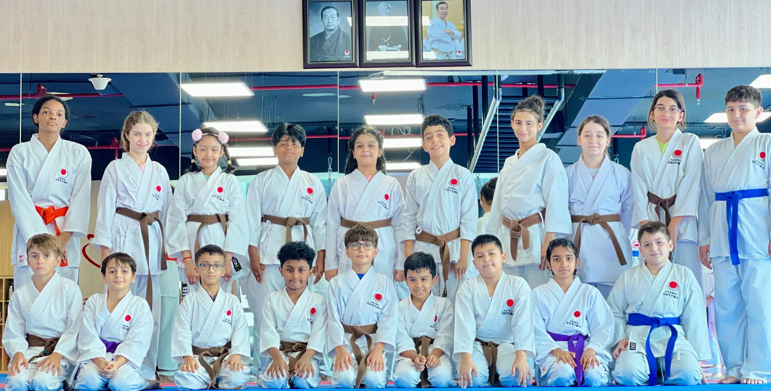 Kids karate class of mixed belt grades posing in rows beneath the dojo's framed portraits