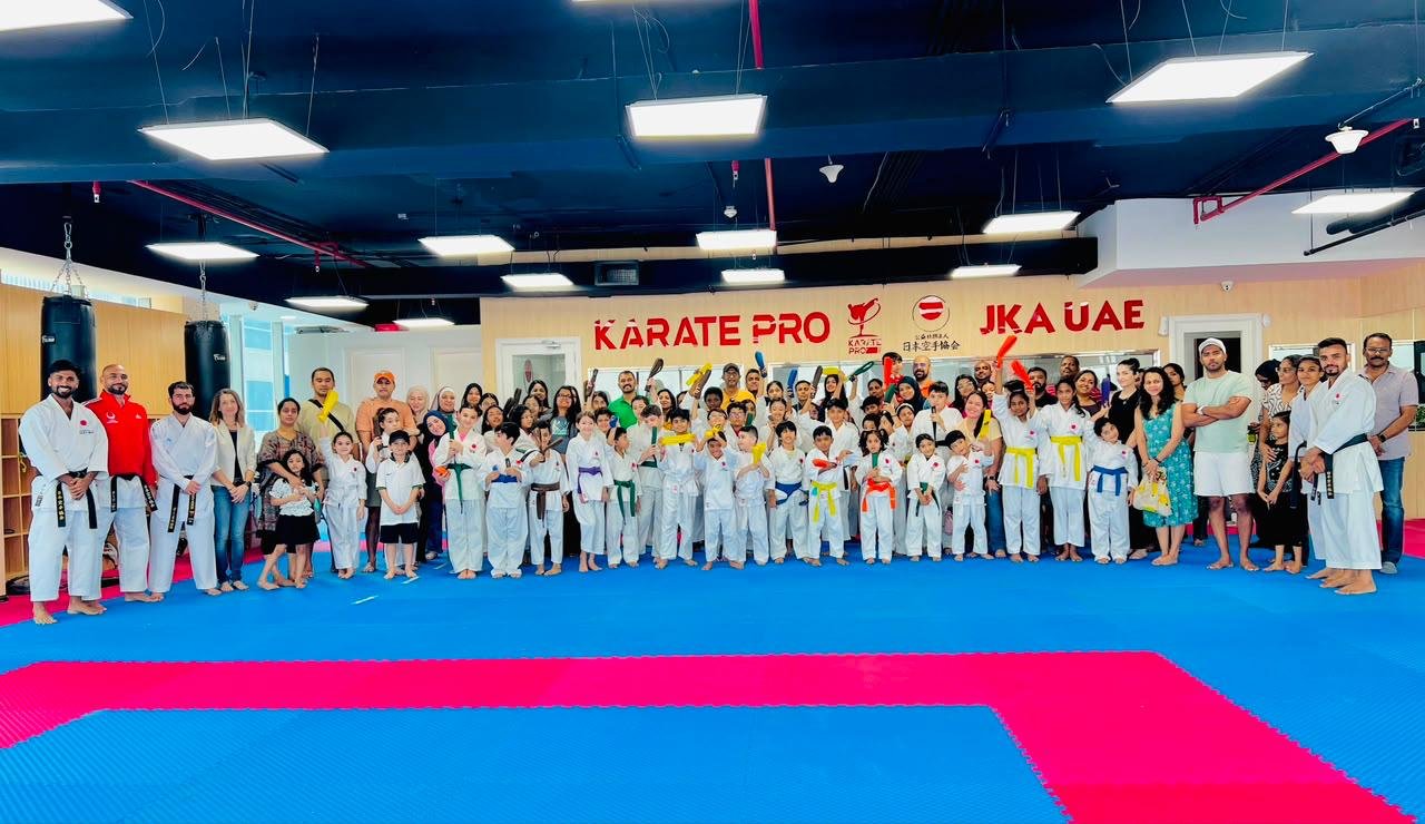 Families and children of mixed belt grades posing together at a belt grading day beneath the Karate Pro and JKA UAE wall signage