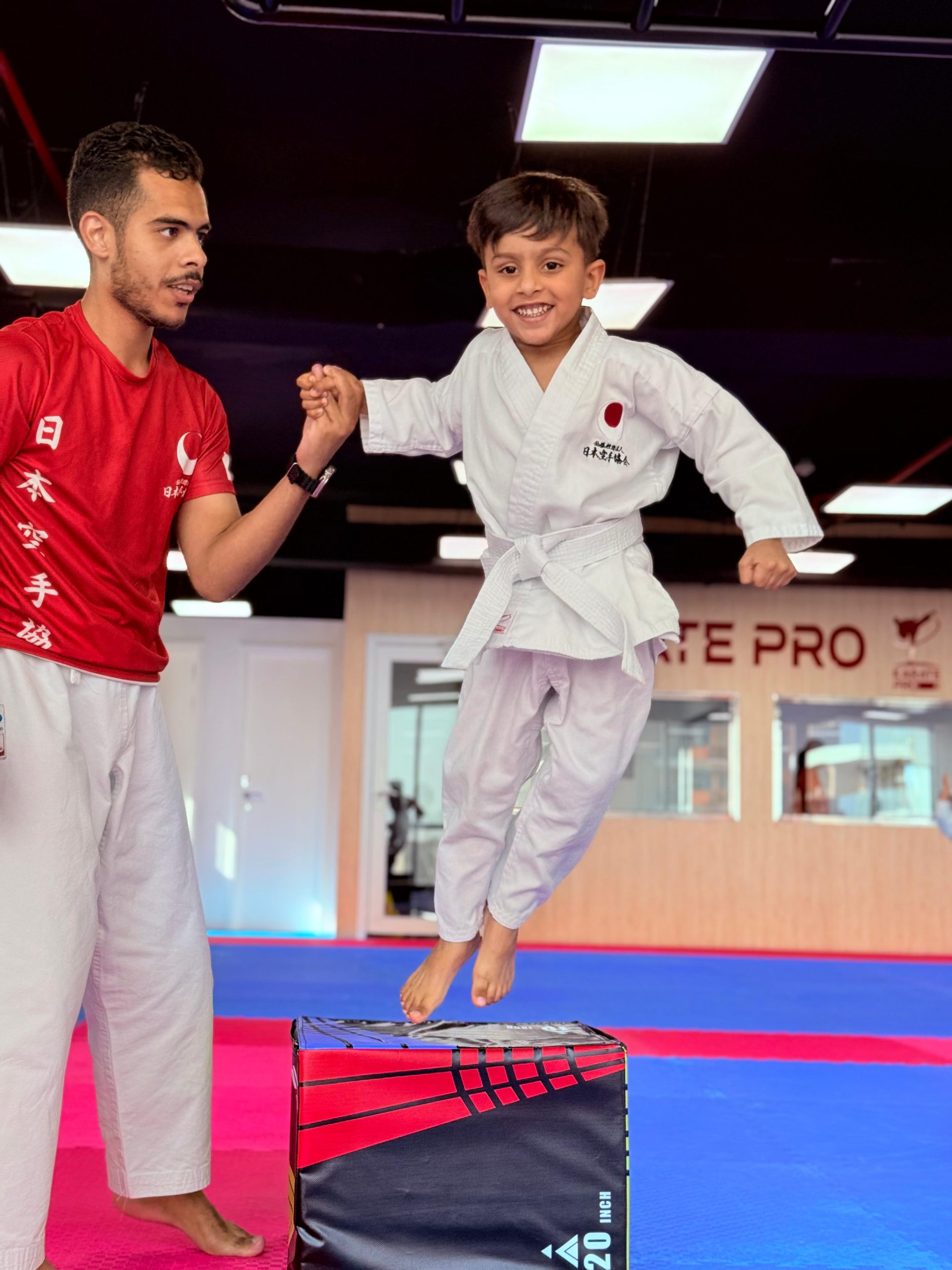 A young boy in white gi jumping off a training box while his coach holds his hand